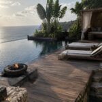 A tropical infinity pool with wooden deck, striped lounge chairs, outdoor shower, and a round stone fire pit set into the edge overlooking the ocean.