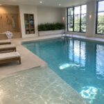 Indoor pool with stone deck, wooden lounge chairs, and a built-in wooden bench with hooks and cushions along the wall.