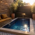 A small rectangular plunge pool with blue mosaic tiles and a brass wall spout pouring water, set in a brick courtyard with patterned tile flooring and potted trees.