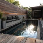 A long rectangular plunge pool with dark water runs between a wooden deck with built-in seating on one side and a gravel path with stone pavers on the other, with large metal planters and a covered seating area in the background.