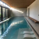 Modern indoor pool with sunlit turquoise water, stone tiles, wooden bench and towels