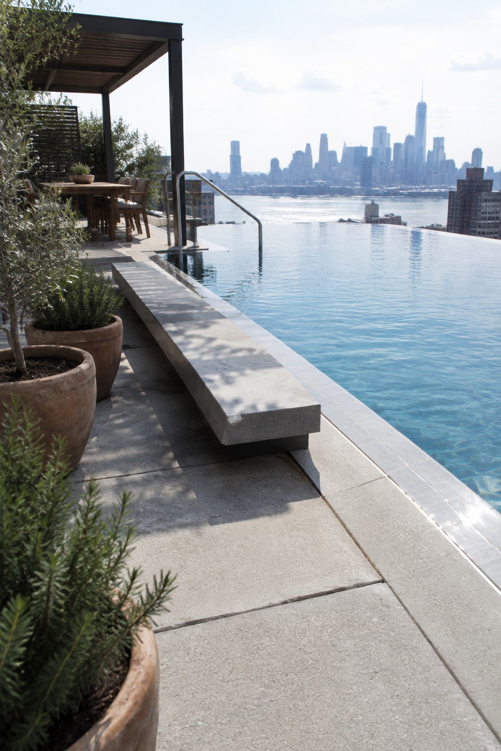 18 Distinctive Infinity Pool Edge Details Featuring Refined Coping Designs An infinity pool with a wide concrete coping ledge that functions as a bench, set on a paved terrace with potted plants and a city skyline in the background.