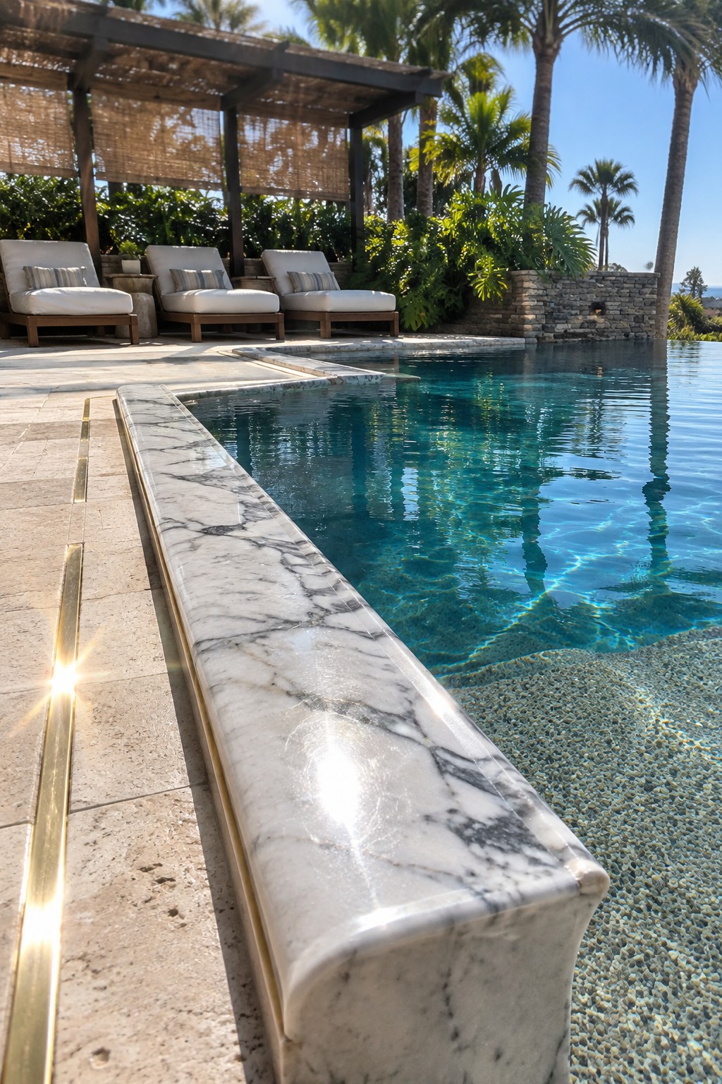 18 Distinctive Infinity Pool Edge Details Featuring Refined Coping Designs A close-up view of an infinity pool edge with white marble coping featuring a thin gold inlay, next to a pebble-bottom section and lounge chairs in the background.