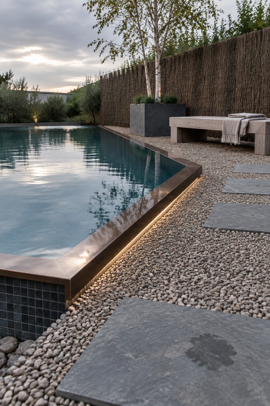 18 Distinctive Infinity Pool Edge Details Featuring Refined Coping Designs A rectangular pool with bronze-toned metal coping and a continuous strip of under-lighting along the edge, bordered by gravel and large stone pavers.
