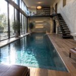 A long indoor lap pool with blue-green tiles in a double-height room with tall windows on the left side and a floating staircase in the background.