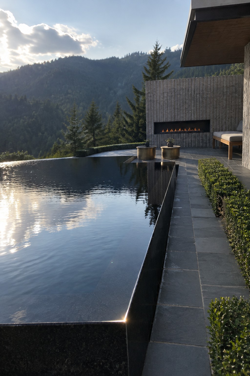 16 Dramatic Infinity Pool Designs With Mountain Views Infinity pool with thick dark stone edging overlooking a mountain range, next to a modern patio with a linear fireplace and round metal tables.