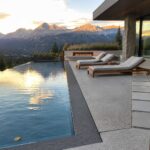 A cantilevered infinity pool with a dark stone edge extends from a concrete patio beside a modern house, with two lounge chairs and mountain peaks visible at sunset.