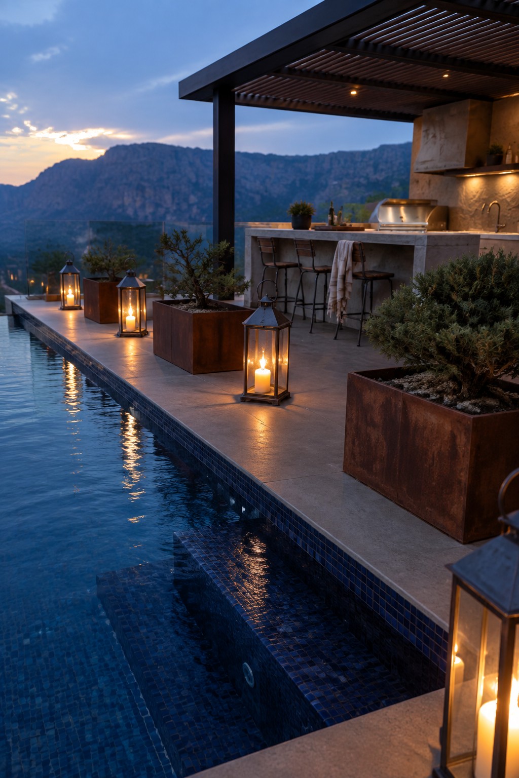 16 Dramatic Infinity Pool Designs With Mountain Views An infinity pool with a concrete outdoor bar, metal planters, and lit lanterns on the deck at dusk overlooking mountains.