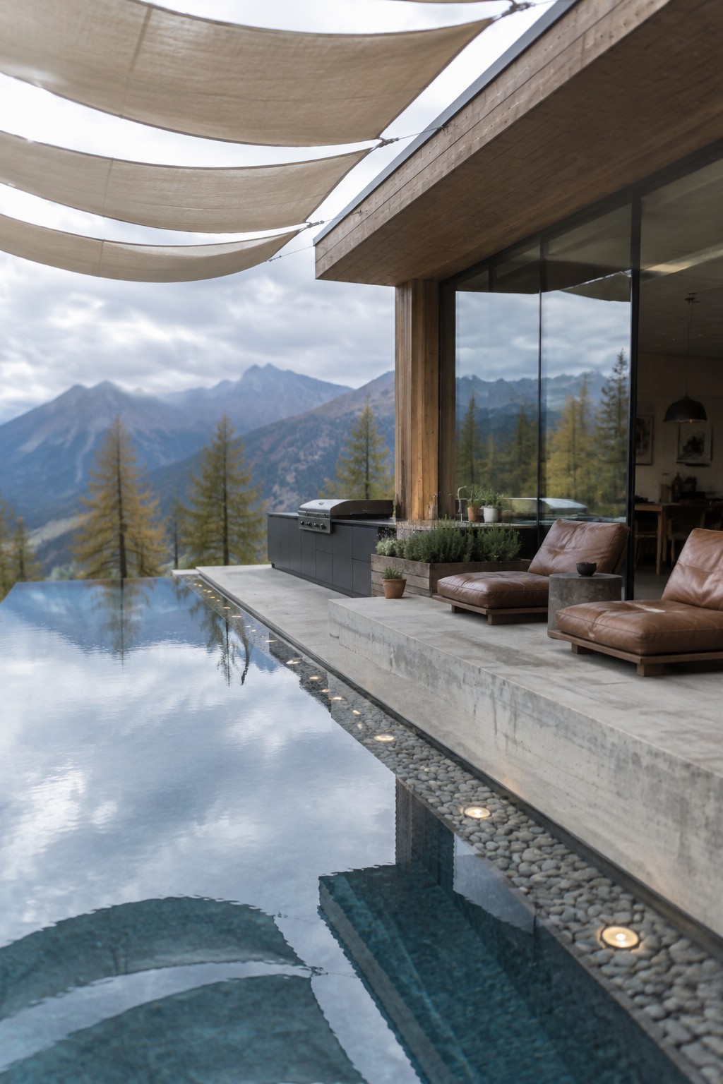 16 Dramatic Infinity Pool Designs With Mountain Views A modern infinity pool with a concrete terrace, brown leather lounge chairs, an outdoor kitchen, and mountains in the background.