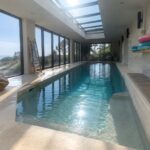 Sunlit indoor lap pool with ocean view, wicker lounges, and pebble wall