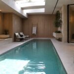 An indoor rectangular pool with turquoise water, a long concrete bench with woven cushions along the left side, vertical wood paneling on the walls, and two skylights in the ceiling.