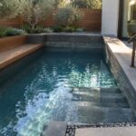 A narrow plunge pool with clear blue water, built-in gray stone steps at one end, and a long wooden bench integrated into the side wall, bordered by pebble paving and trees.