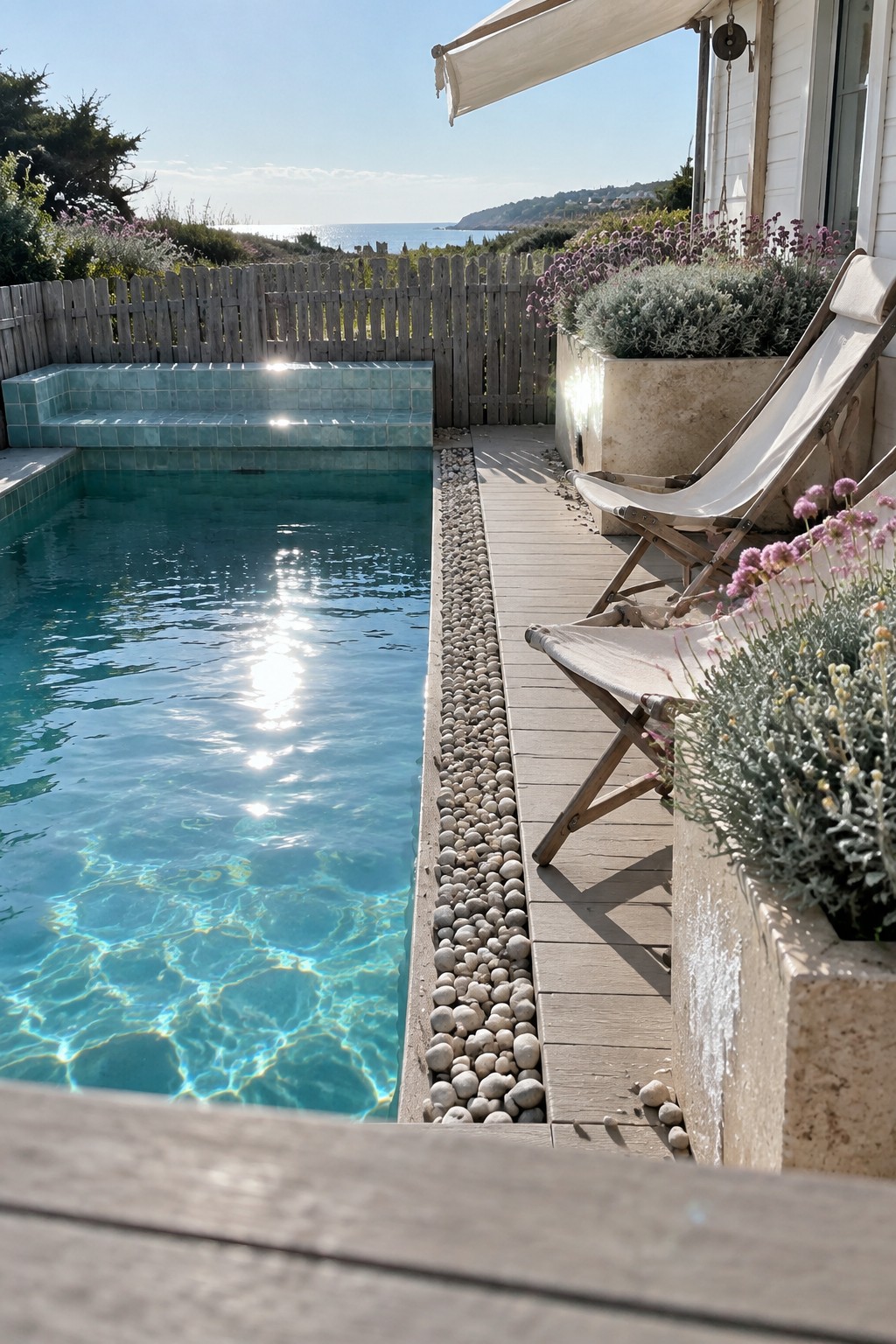 A narrow plunge pool with clear blue water sits beside a strip of light pebbles that separates it from a wooden deck holding two canvas lounge chairs and large potted plants.
