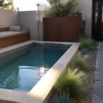 A long rectangular plunge pool with light stone coping, surrounded by gravel beds holding clumps of tall ornamental grasses and a wooden bench with cushions along one side.