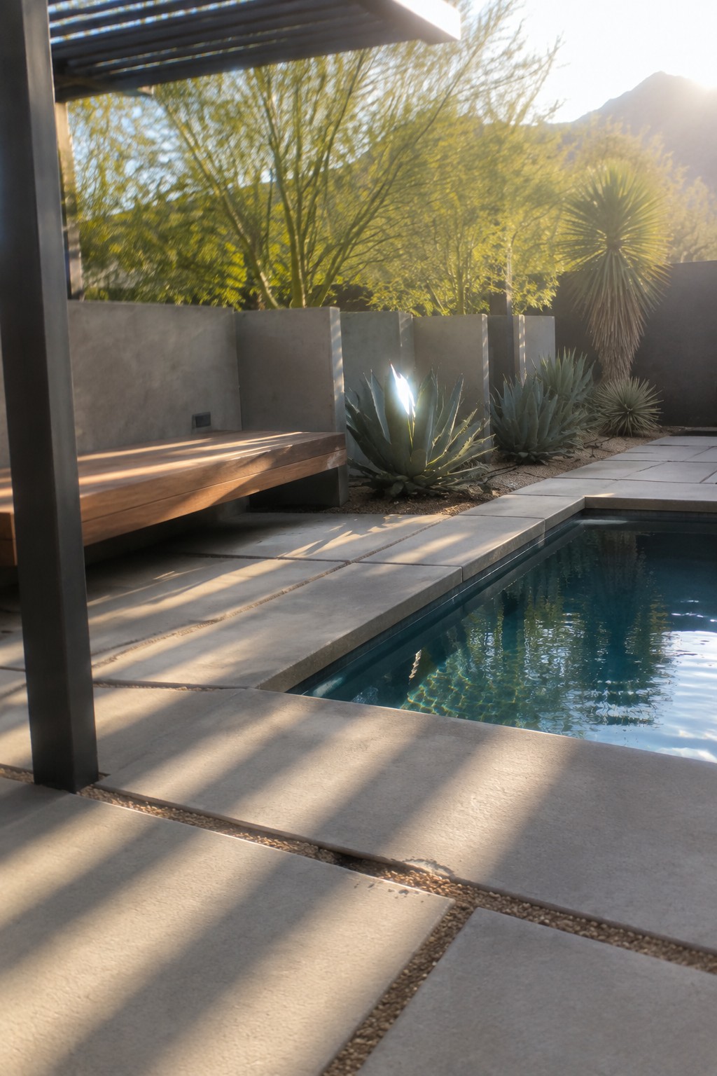 A plunge pool edged with large concrete pavers, a wooden bench set into gray concrete walls, and a few agave plants nearby.