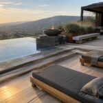 A modern backyard infinity pool with wide wooden decking, two dark gray lounge chairs in the foreground, and a fire pit plus covered seating area overlooking hills at sunset.