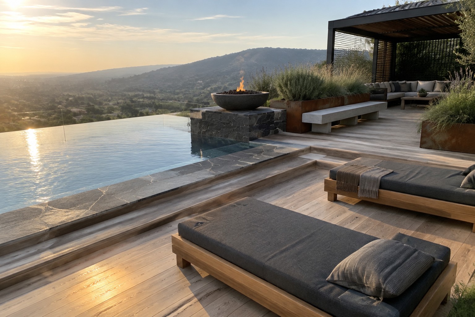 18 Stunning Backyard Infinity Pool Ideas With Modern Outdoor Layouts A modern backyard infinity pool with wide wooden decking, two dark gray lounge chairs in the foreground, and a fire pit plus covered seating area overlooking hills at sunset.