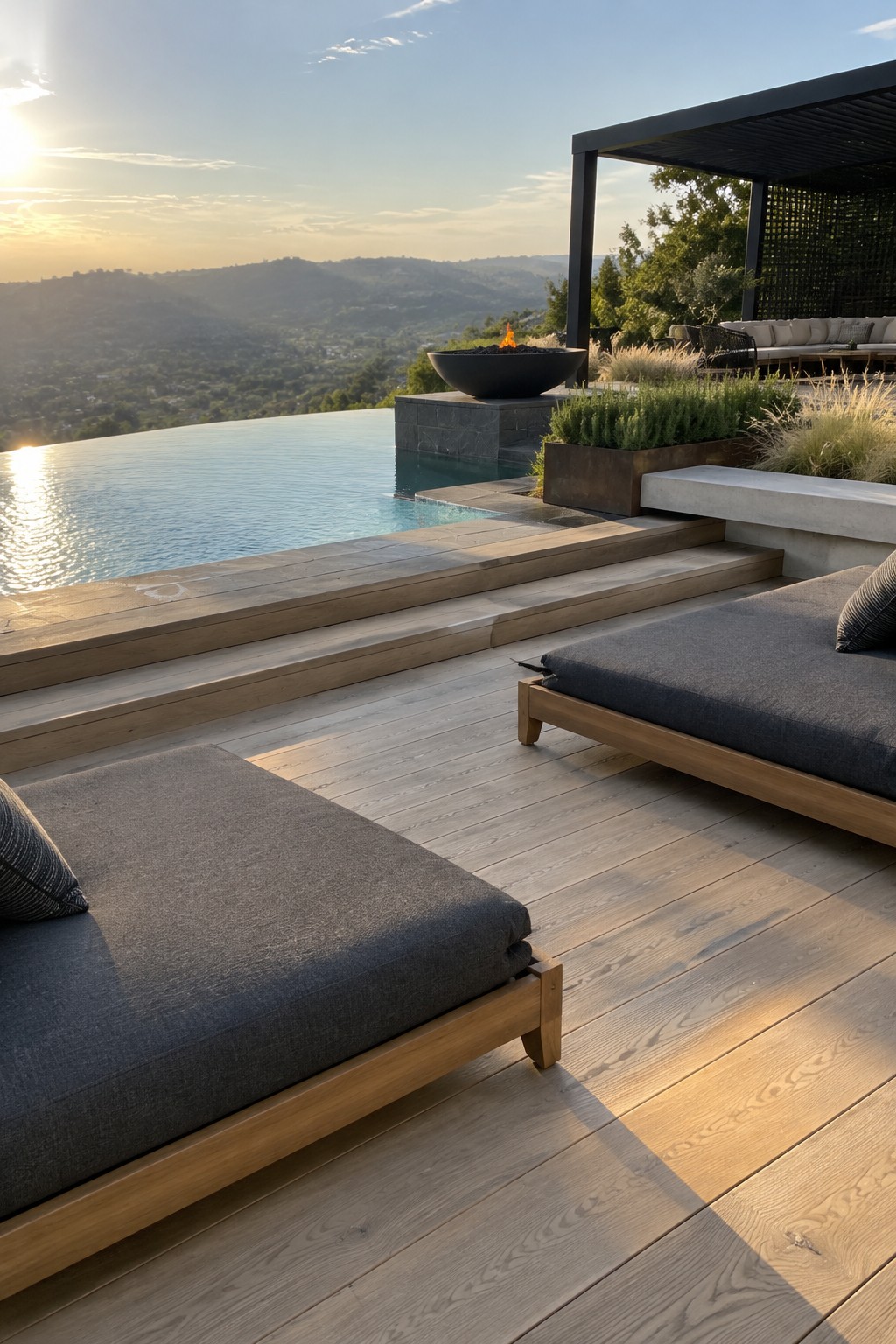 18 Stunning Backyard Infinity Pool Ideas With Modern Outdoor Layouts A modern backyard infinity pool with wide wooden decking, two dark gray lounge chairs in the foreground, and a fire pit plus covered seating area overlooking hills at sunset.
