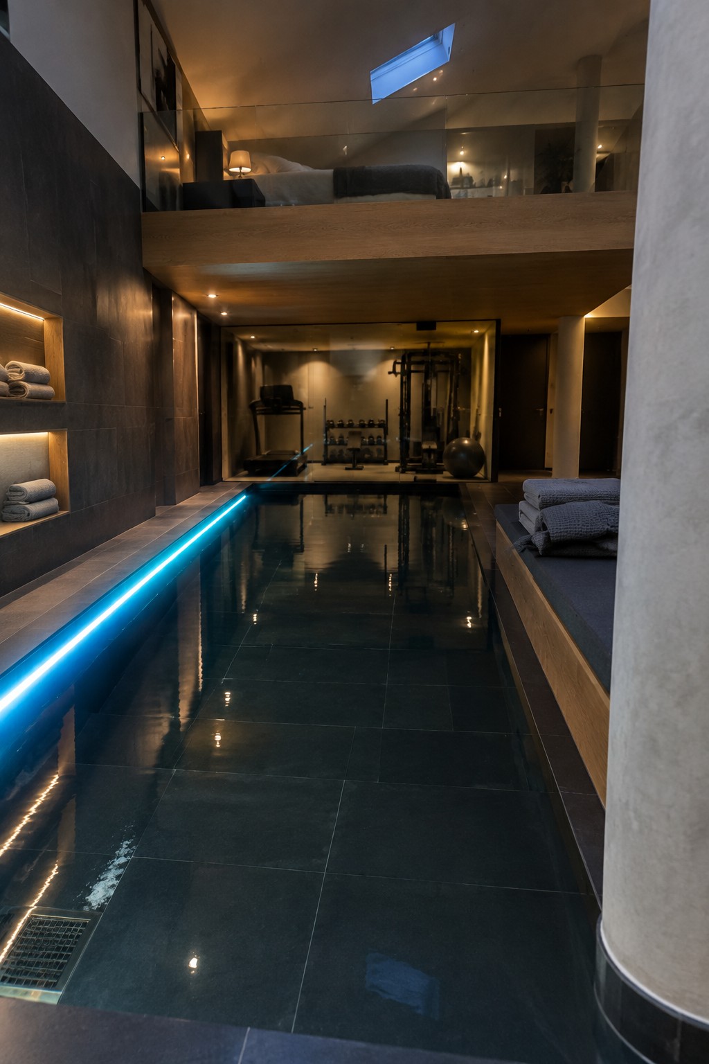 A narrow indoor lap pool with a bright blue LED strip along the left edge, dark tile decking, built-in towel niches on the left wall, and a gym area visible at the far end beneath a glass-railed loft bedroom.