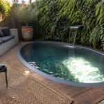 An elongated oval plunge pool with a wall-mounted waterfall sits on a wooden deck beside a built-in concrete bench with gray cushions.