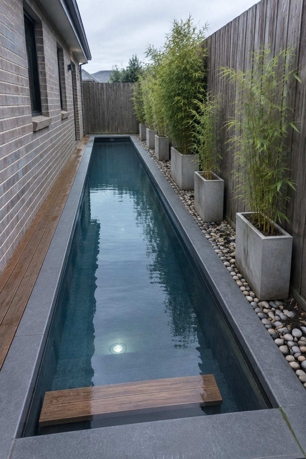 A long narrow plunge pool sits between a brick house wall and a wooden fence, with concrete planters holding bamboo and a wooden bench inside the water at one end.