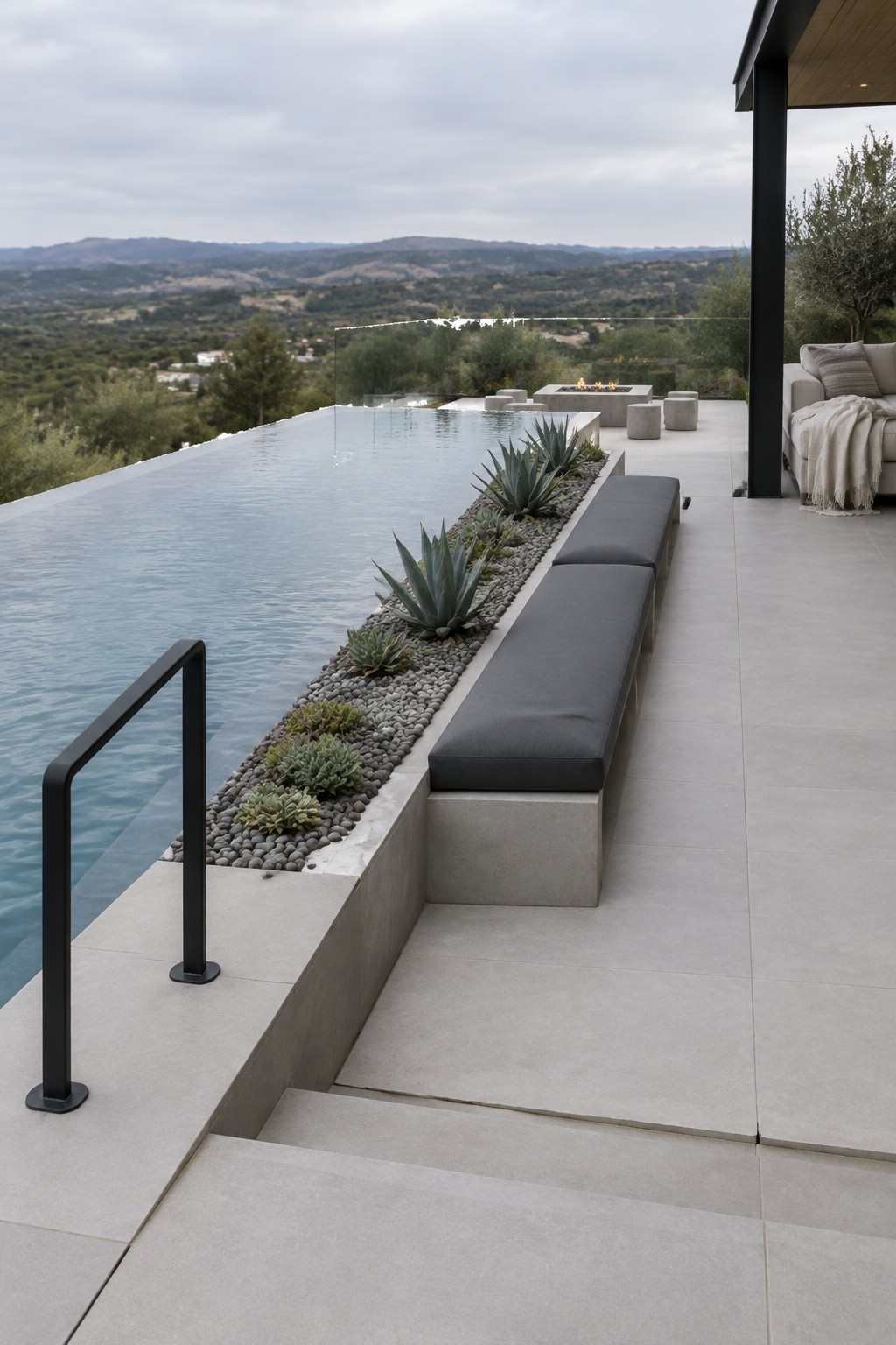 An infinity pool with a long built-in concrete bench with dark gray cushions, a gravel bed with succulents between the bench and the water, and a fire pit area in the background on a tiled patio.