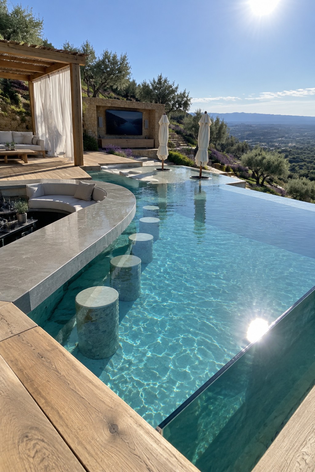 An infinity pool with a curved built-in seating ledge, submerged stone stools, a bar counter, and an outdoor lounge area with a television mounted on a stone wall.