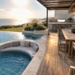 An infinity pool with curved built-in seating and cushions sits beside a wooden deck with a dining table, bar stools, and outdoor kitchen overlooking the ocean.