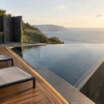 An angular infinity pool with a glass barrier borders a wooden deck with two lounge chairs and an outdoor shower, overlooking the ocean.