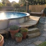 Above ground pool with pallet wood decking and steps, potted plants, gravel path with stone edging, and string lights in a backyard garden.