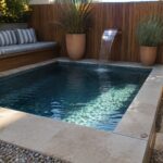 A rectangular plunge pool set in a corner with built-in wooden bench seating along two walls, striped cushions, a copper water spout on the left, and large terracotta pots nearby.