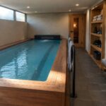 An indoor endless pool with teal water, surrounded by a light wood deck and bench, with wall shelves holding towels and a sauna door on the right side of the room.
