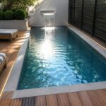 A rectangular plunge pool with a wall-mounted waterfall, bordered by wooden decking, striped lounge chairs, and a concrete planter with shrubs.