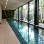 An indoor lap pool runs alongside floor-to-ceiling glass walls with black frames, showing trees and greenery outside, while a green living wall stands at the far end and light wood flooring extends along the right side.