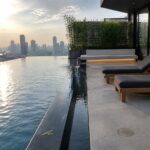 An infinity pool with a narrow linear water channel along its edge, two dark lounge chairs on a concrete terrace, and a city skyline in the background.