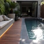 A narrow plunge pool runs parallel to a long built-in concrete bench with light cushions on a wooden deck, bordered by potted plants and a living wall.