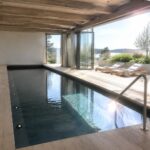 An indoor pool with dark edging runs alongside light wood flooring, with large glass doors open to an outdoor patio holding two lounge chairs and distant greenery.