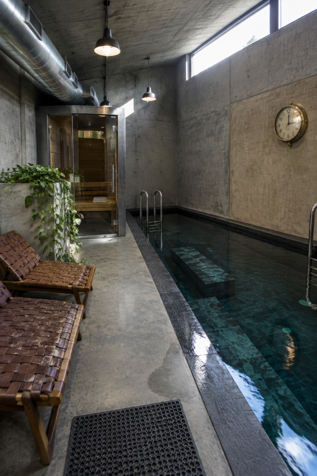An indoor lap pool with dark water beside a glass sauna enclosure, concrete walls, woven lounge chairs, and a round wall clock.