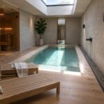 Modern indoor pool with wooden lounge chairs, sauna, skylight, and spa decor.