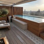 A raised rectangular plunge pool with light blue water is set into a wooden deck, with a concrete bench running along one side and two cushioned lounge chairs positioned nearby.