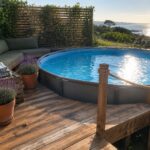 An above-ground pool on a wooden deck with rope railings between posts, a small wooden ladder, potted lavender plants, and built-in seating with green cushions.