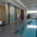Modern indoor lap pool with turquoise water, gray tiles, wood lockers and wall clock.