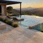 An outdoor infinity pool with blue-green glass mosaic tiles along the edge, two lounge chairs partially in the water, a stone fire pit, and a hillside landscape at sunset.