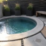 A round plunge pool with a wall-mounted water spout sits in a concrete patio area bordered by black planters, a wooden bench, and pebble edging.