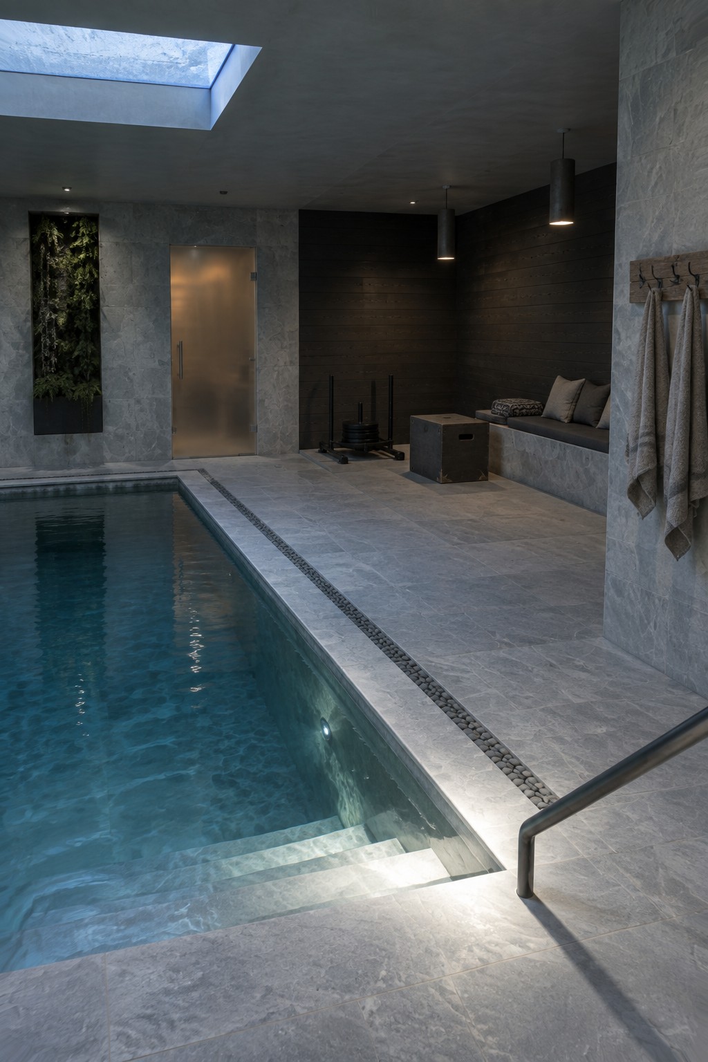 Modern indoor pool with stone tiles, wooden walls, skylight, and hanging towels in spa.