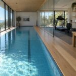 Modern indoor lap pool with blue tiles, sunlight reflections, wooden floors, and gym area.