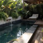 Sunlit pool with waterfall, rattan loungers on wooden deck amid tropical plants