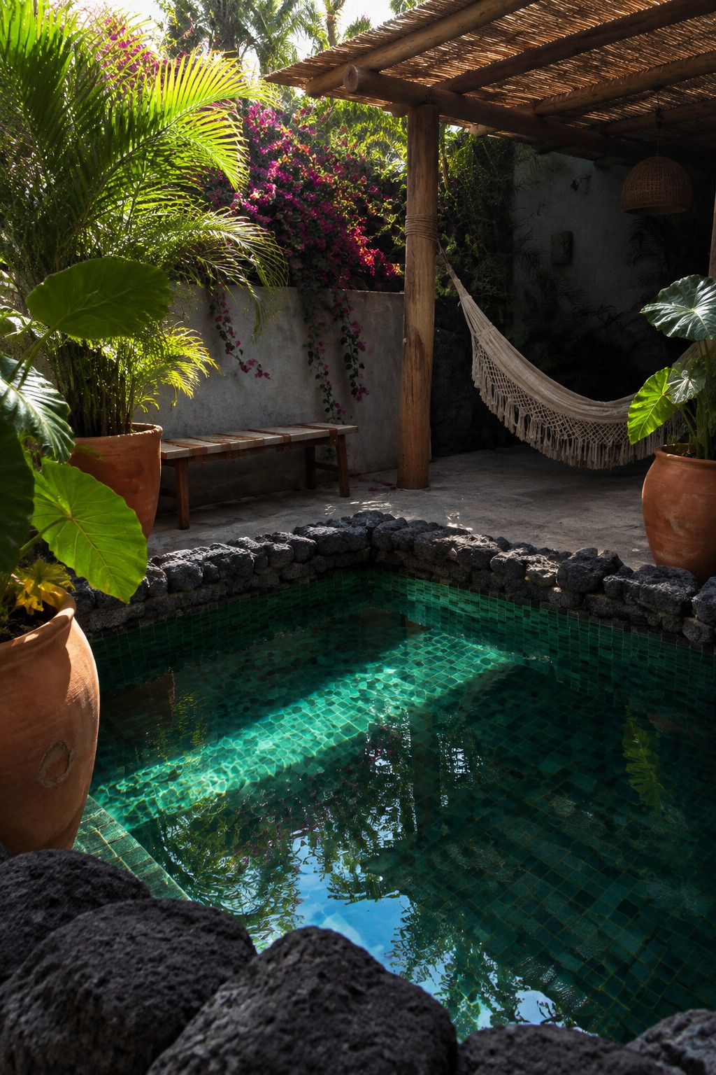 Emerald-tiled pool ringed by dark rocks in sunny tropical garden with hammock