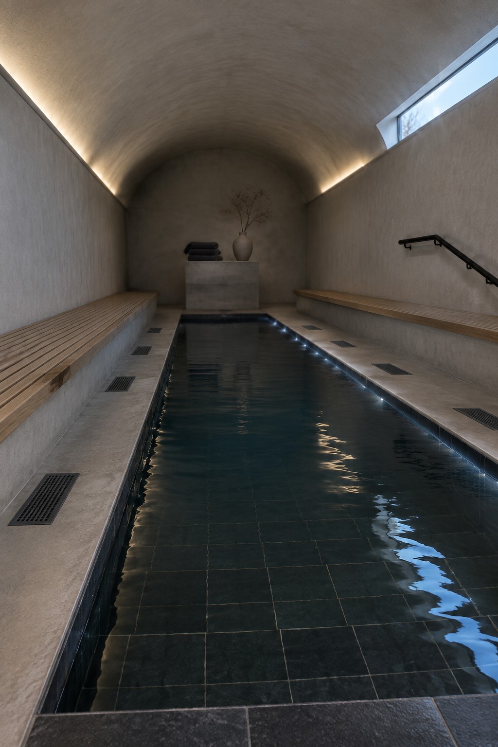 Minimalist vaulted spa with long dark-tiled pool, wooden benches, and stone pedestal.