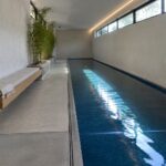 A long narrow indoor pool with blue mosaic tiles runs through a minimalist concrete space, with a wooden bench and potted plant on the left and a recessed niche holding rolled towels on the right.